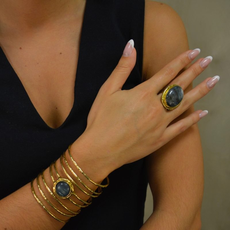 la manchette Kiera en laiton doré à l'or fin incarne l’élégance moderne avec une pierre de labradorite ronde, resplendissante au centre. Sa silhouette large et ajourée offre un mélange audacieux de sophistication et d’originalité. Bijou porté, Crédit GEMITY.