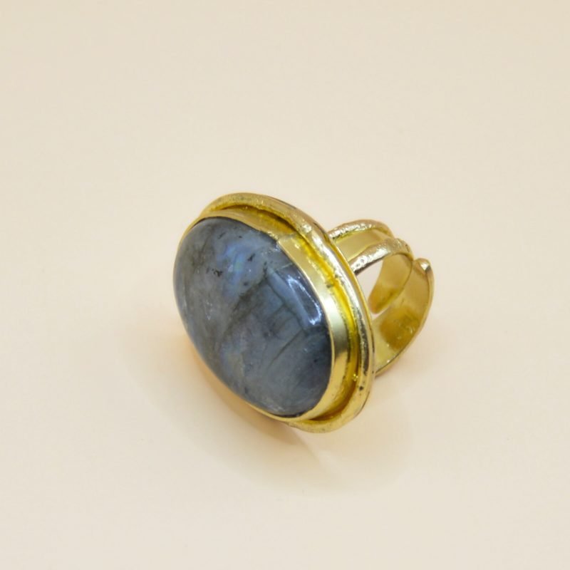 Confectionnée avec soin en laiton doré à l'or fin 18 carats, la bague Mystique est réglable et capture l'essence de l'inconnu et de la fascination. En son cœur, un imposant cabochon de labradorite scelle l'alliance entre l'éclat doré et la profondeur changeante de la pierre. Bijou seul, Crédit GEMITY