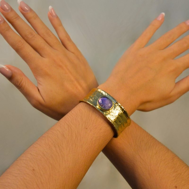 Bracelet jonc ajustable inspiré des mystères du désert. Son design et sa texture lisse rappelle les subtiles nuances du sable doré qui s'étend à perte de vue, tandis que sa pierre centrale en améthyste ajoute une touche d'élégance et de fraîcheur. . Bijou porté, Crédit GEMITY.