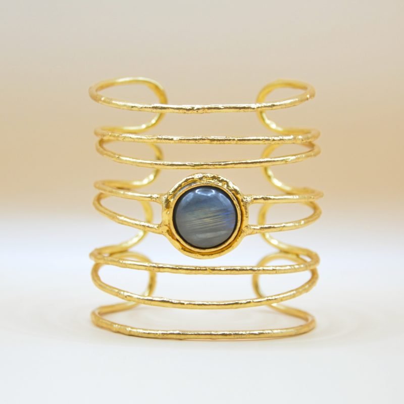 la manchette Kiera en laiton doré à l'or fin incarne l’élégance moderne avec une pierre de labradorite ronde, resplendissante au centre. Sa silhouette large et ajourée offre un mélange audacieux de sophistication et d’originalité. Bijou seul, Crédit GEMITY