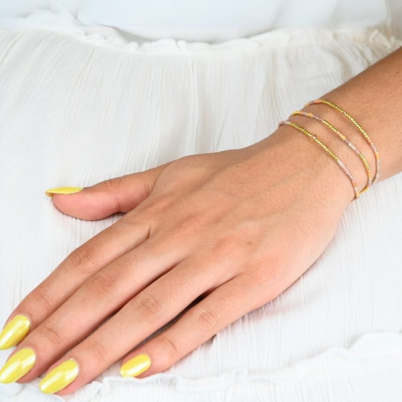 ce bracelet fin à 3 rangs combine des perles dorées, nude et dans les tons rosés pour un design doux et féminin. L'un des rangs est orné d'une plus grande perle au centre, apportant une touche de sophistication et de brillance. . Bijou porté, Crédit GEMITY.