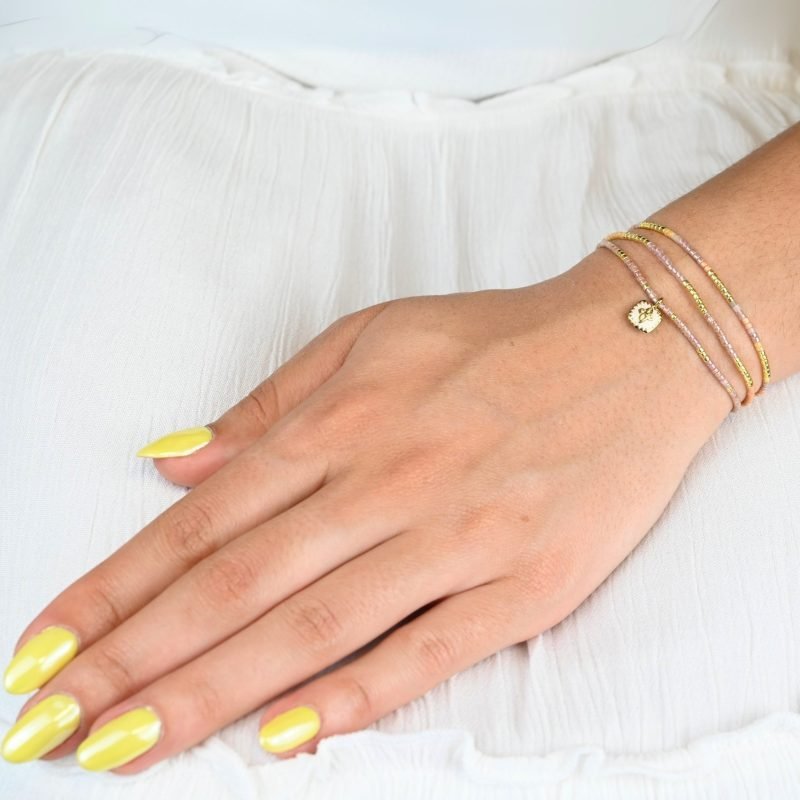 ce bracelet fin à 3 rangs combine des perles dorées et nude pour un effet élégant et doux. Il est orné d'un charm fleur doré, posé sur un fond en nacre blanc en forme de carré, apportant une touche de sophistication et de brillance. . Bijou porté, Crédit GEMITY.