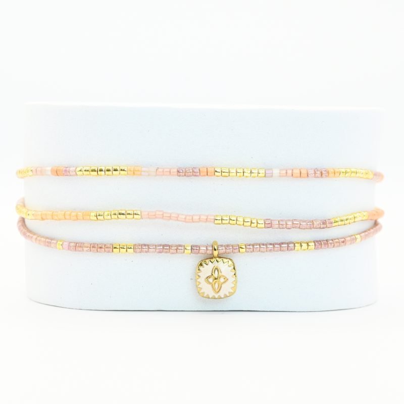 ce bracelet fin à 3 rangs combine des perles dorées et nude pour un effet élégant et doux. Il est orné d'un charm fleur doré, posé sur un fond en nacre blanc en forme de carré, apportant une touche de sophistication et de brillance. . Bijou seul, Crédit GEMITY