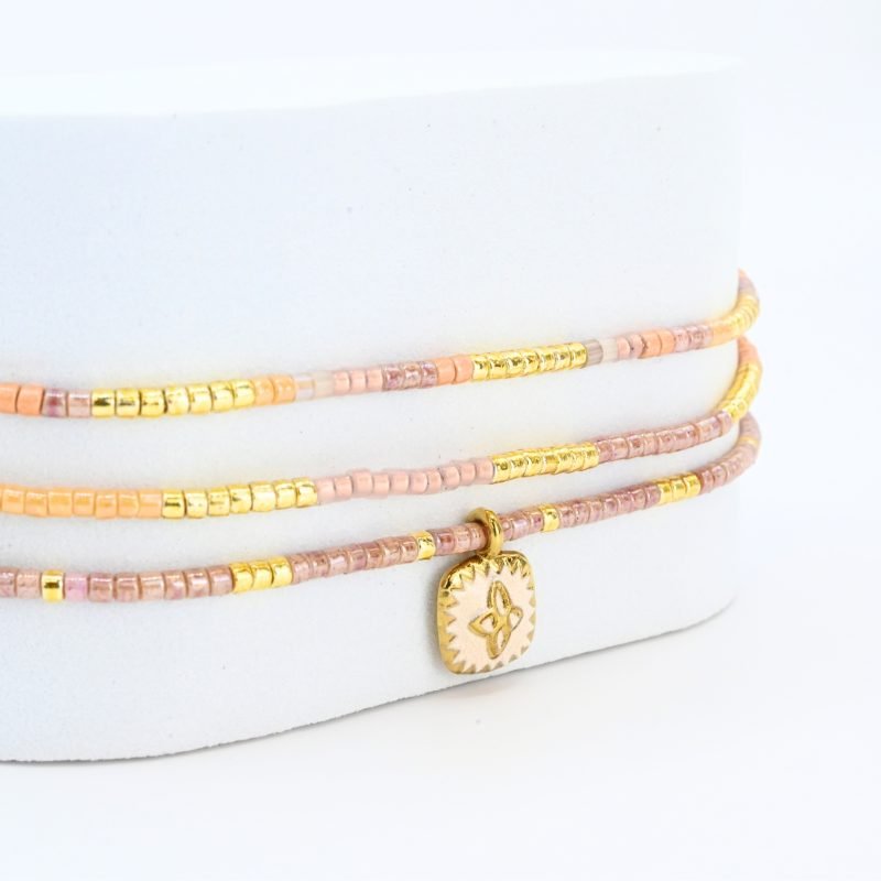 ce bracelet fin à 3 rangs combine des perles dorées et nude pour un effet élégant et doux. Il est orné d'un charm fleur doré, posé sur un fond en nacre blanc en forme de carré, apportant une touche de sophistication et de brillance. . Bijou seul, Crédit GEMITY