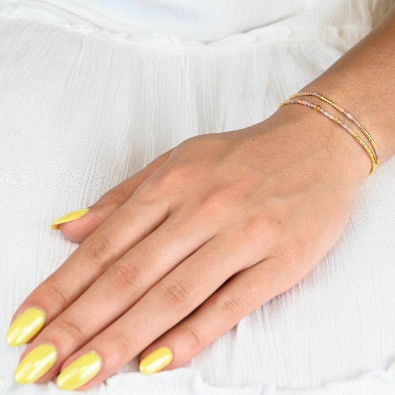 ce bracelet fin à 2 rangs associe des perles dorées, blanches, roses et nude pour un design doux et coloré. L'un des rangs est orné d’une perle plus grosse au centre, apportant une touche de sophistication à ce bijou délicat. . Bijou porté, Crédit GEMITY.