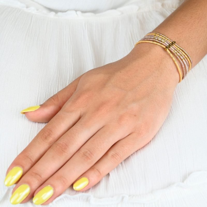 ce bracelet fin à 6 rangs associe des perles dorées et nude pour un look élégant et raffiné. Un ornement traverse les six rangs, créant un effet unique et structuré. . Bijou porté, Crédit GEMITY.