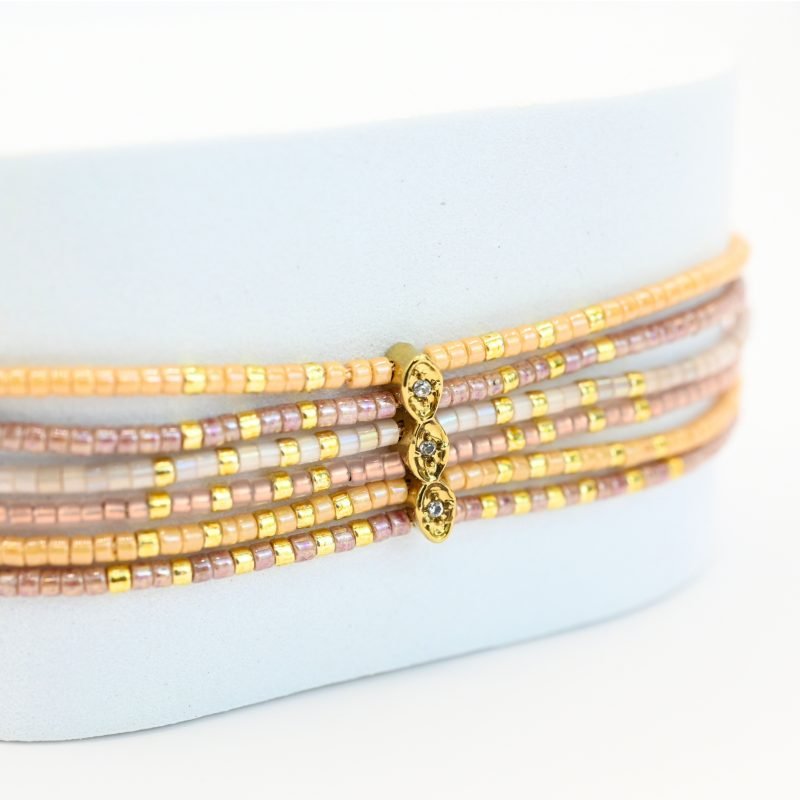 ce bracelet fin à 6 rangs associe des perles dorées et nude pour un look élégant et raffiné. Un ornement traverse les six rangs, créant un effet unique et structuré. . Bijou seul, Crédit GEMITY