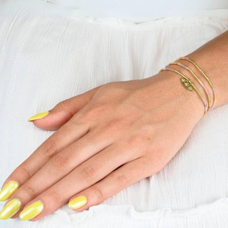 ce bracelet à 3 rangs combine des perles dorées, nude et rose poudré. Le second rang est orné d’un charm œil au centre, ajoutant une touche originale et élégante. . Bijou porté, Crédit GEMITY.