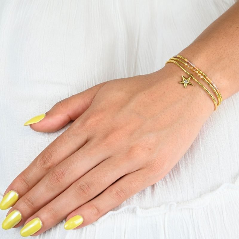 ce bracelet raffiné à 3 rangs combine des perles dorées et nude pour un look élégant et féminin. Le premier rang est orné de perles délicates, tandis que le second présente des perles plus grosses, créant un effet visuel dynamique. Le troisième rang se distingue par un charm étoile au centre, ajoutant une touche de magie et de sophistication. . Bijou porté, Crédit GEMITY.
