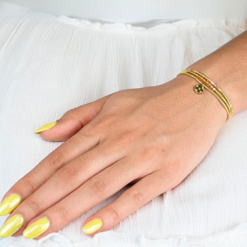 ce bracelet fin à 3 rangs combine des perles dorées, nude et roses, créant une harmonie de couleurs douce et élégante. Le charm rond à strass, placé au centre, ajoute une touche scintillante et raffinée. . Bijou porté, Crédit GEMITY.