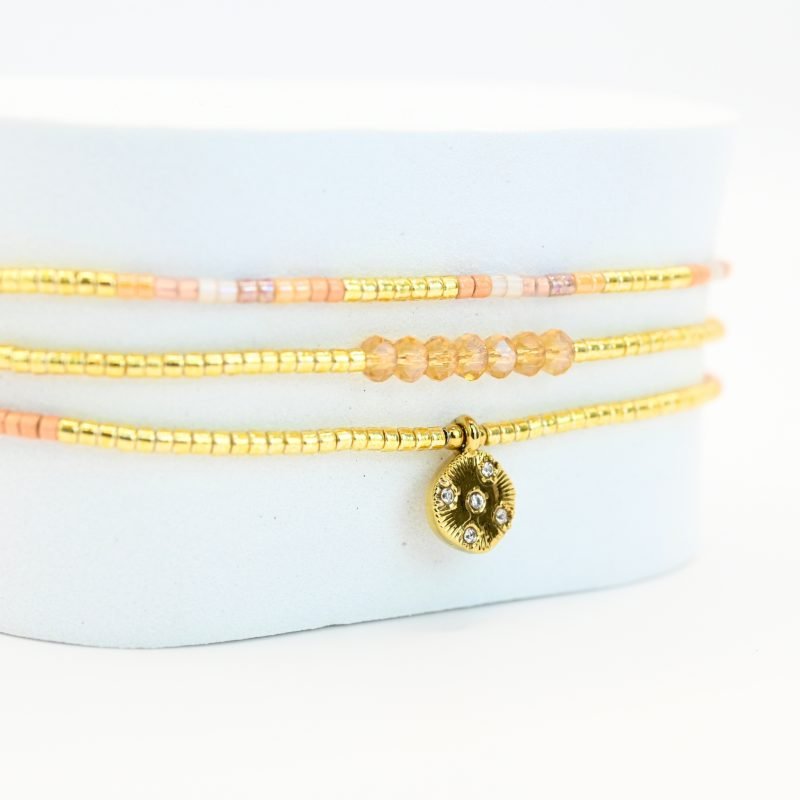 ce bracelet fin à 3 rangs combine des perles dorées, nude et roses, créant une harmonie de couleurs douce et élégante. Le charm rond à strass, placé au centre, ajoute une touche scintillante et raffinée. . Bijou seul, Crédit GEMITY