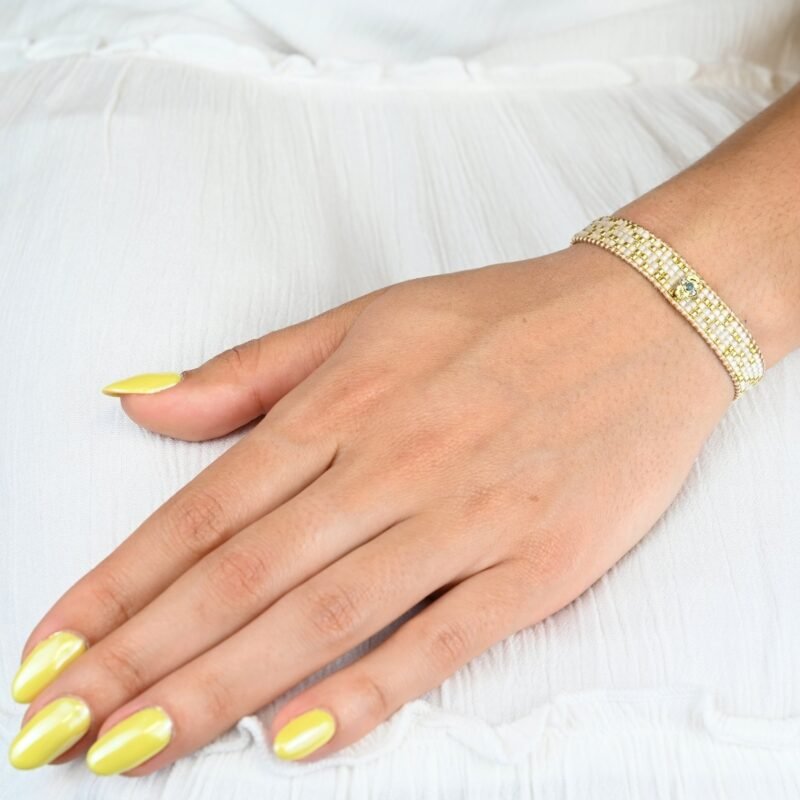 ajoutez une touche de sophistication à votre poignet avec cette petite manchette composée de 5 rangs de perles dorées et blanches. L'ornement doré en son centre apporte une touche de brillance et d'élégance supplémentaire à ce bijou délicat.. Bijou porté, Crédit GEMITY.