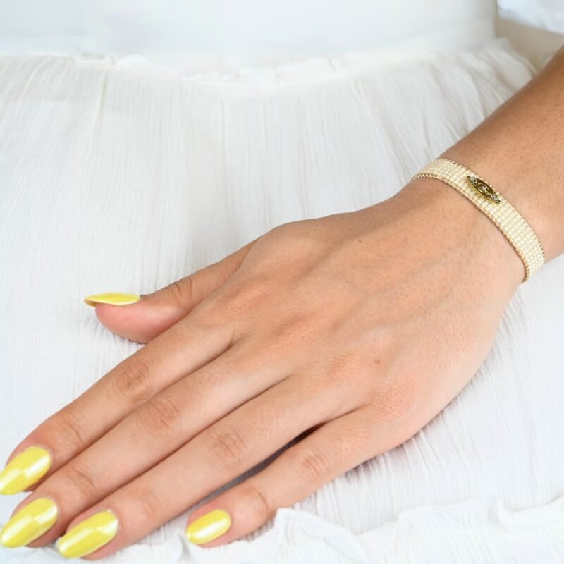 cette petite manchette raffinée se compose de 5 rangs de perles blanches, offrant une touche lumineuse et élégante. Son ornement central en forme d'œil apporte une symbolique protectrice et tendance. . Bijou porté, Crédit GEMITY.
