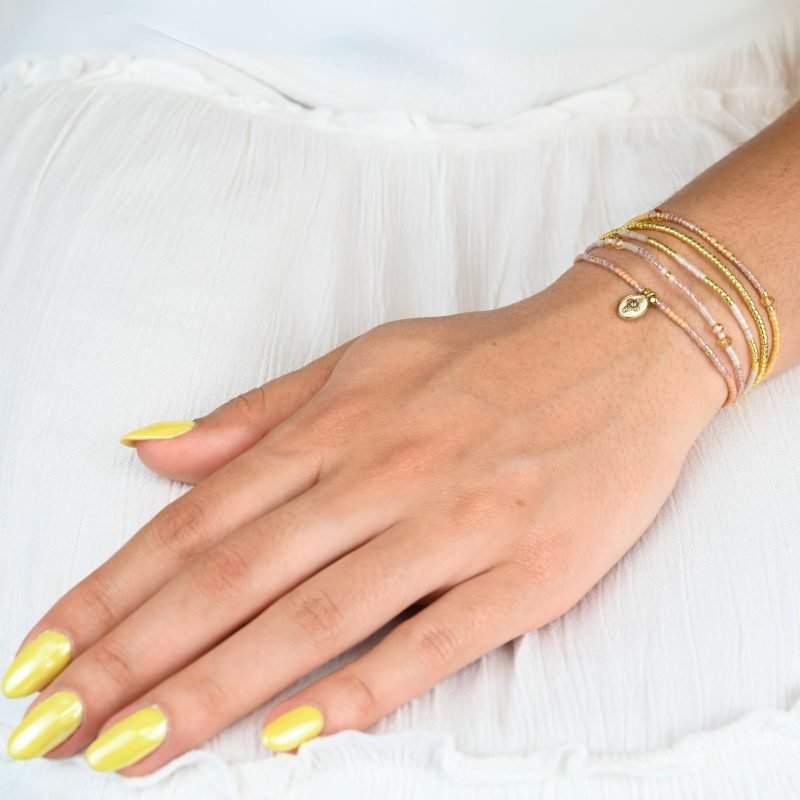 ce bracelet fin à 5 rangs combine des perles dorées, nude, blanches et dans les tons rosés, créant un design doux et harmonieux. Un charm fleur doré, avec un zircon au centre, est placé sur un fond blanc en forme ovale sur l’un des rangs, ajoutant une touche élégante et raffinée. . Bijou porté, Crédit GEMITY.