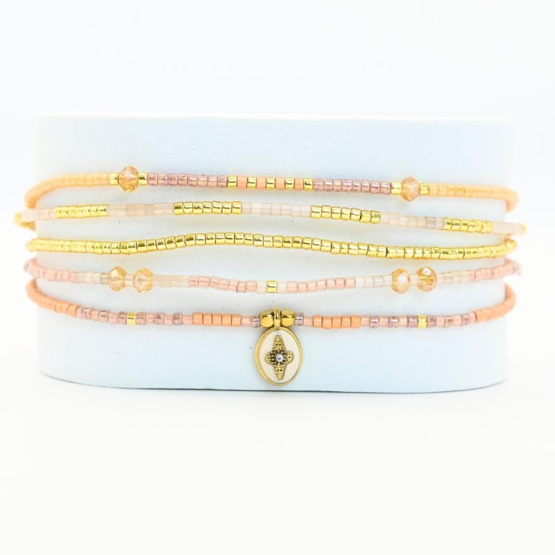 ce bracelet fin à 5 rangs combine des perles dorées, nude, blanches et dans les tons rosés, créant un design doux et harmonieux. Un charm fleur doré, avec un zircon au centre, est placé sur un fond blanc en forme ovale sur l’un des rangs, ajoutant une touche élégante et raffinée. . Bijou seul, Crédit GEMITY