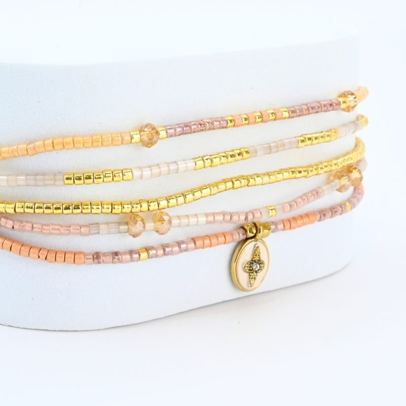 ce bracelet fin à 5 rangs combine des perles dorées, nude, blanches et dans les tons rosés, créant un design doux et harmonieux. Un charm fleur doré, avec un zircon au centre, est placé sur un fond blanc en forme ovale sur l’un des rangs, ajoutant une touche élégante et raffinée. . Bijou seul, Crédit GEMITY