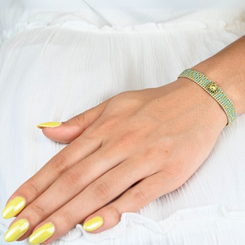 sublimez votre poignet avec cette petite manchette délicate, composée de 5 rangs de perles dorées et vert d'eau qui apportent une touche fraîche et lumineuse. L'ornement soleil au centre ajoute une note rayonnante et éclatante, idéale pour apporter une touche d'élégance à vos tenues.. Bijou porté, Crédit GEMITY.