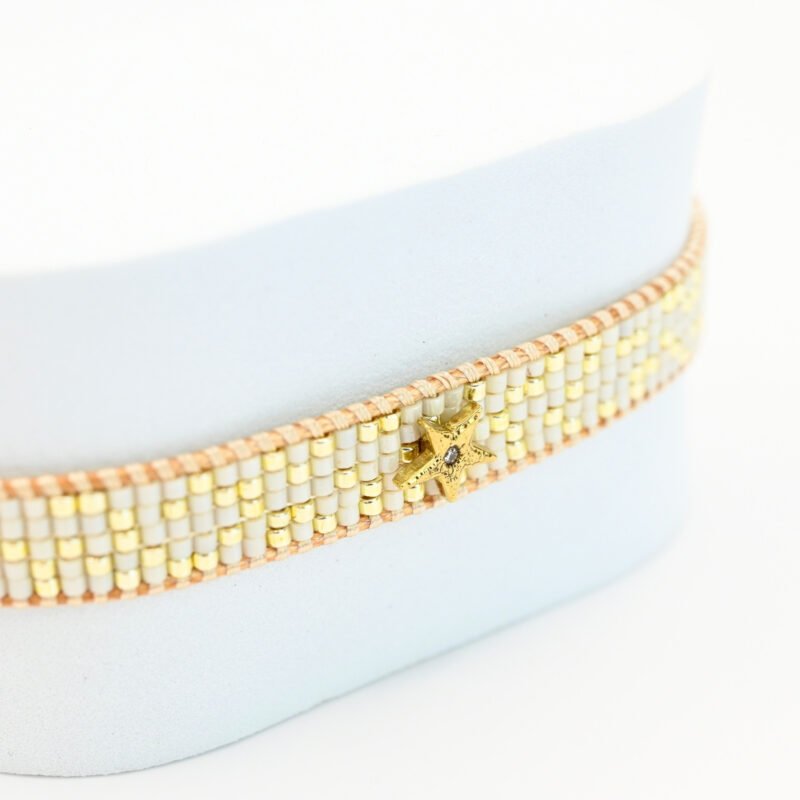 cette petite manchette allie délicatesse et élégance grâce à ses 5 rangs de perles blanches et dorées, créant un contraste subtil et raffiné. L'ornement étoilé en son centre ajoute une touche de brillance et de magie à ce bijou.. Bijou posé, Crédit GEMITY.