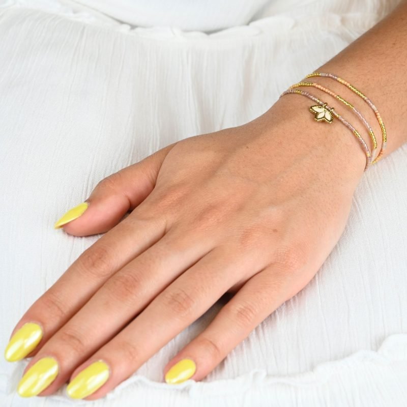ce bracelet fin à 3 rangs associe des perles dorées, nude et dans les tons rosés pour un effet doux et élégant. Il est orné d’un charm fleur de lotus en nacre, posé sur un fond doré de la même forme, apportant une touche de raffinement et de luminosité. . Bijou porté, Crédit GEMITY.