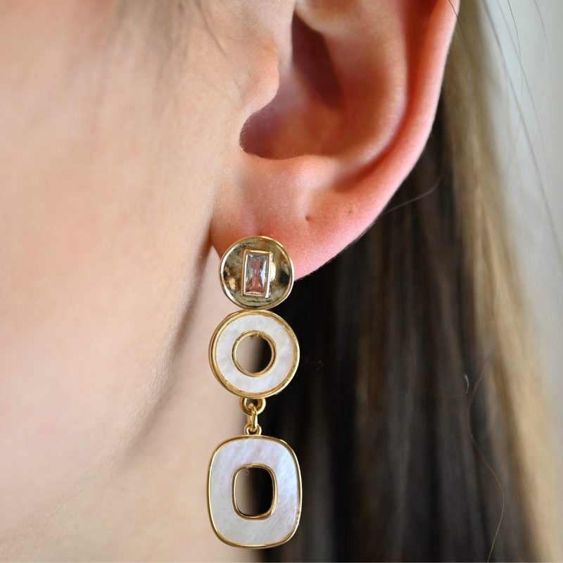 "Boucles d’oreilles à tige en métal doré à l’or fin et pendant en métal doré et nacres naturelles. Ces charmantes boucles d’oreilles fantaisie en plaqué or sont sont un excellent ajout à tout autre bijoux. Elles ne provoquent pas d'allergies au nickel. Taille : 6 cm . Bijou porté, Crédit GEMITY."
