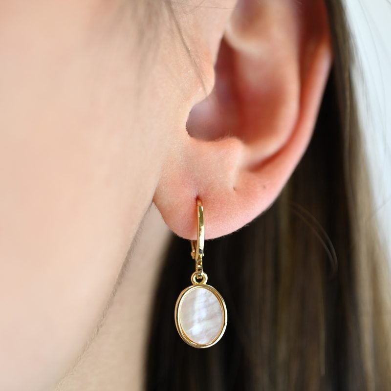 "Boucles d’oreilles dormeuses avec une nacre découpée en ovale entourée par le métal doré à l’or fin. La nacre naturelle est mise en valeur de manière subtile mais envoûtante, avec ses irisations délicates qui scintillent à la lumière. Taille du pendant : 12 mm. Bijou porté, Crédit GEMITY."