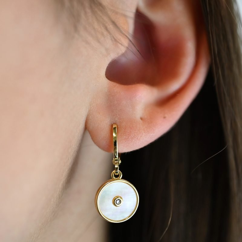 Boucles d'oreilles en métal et cabochon en nacre naturelle de 1 cm. Une nacre naturelle qui se portera avec délicatesse contre votre peau. Elle est composée d’un petit pendentif avec en son centre un zirconium couleur cristal. . Bijou porté, Crédit GEMITY.