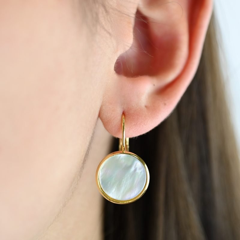 "Boucles d’oreilles dormeuses et cabochon en nacre de 14 mm. Elles sont dorées à l’or fin 24 carats. Ces boucles apporteront une touche lumineuse à vos tenues dans un style épuré. . Bijou porté, Crédit GEMITY."