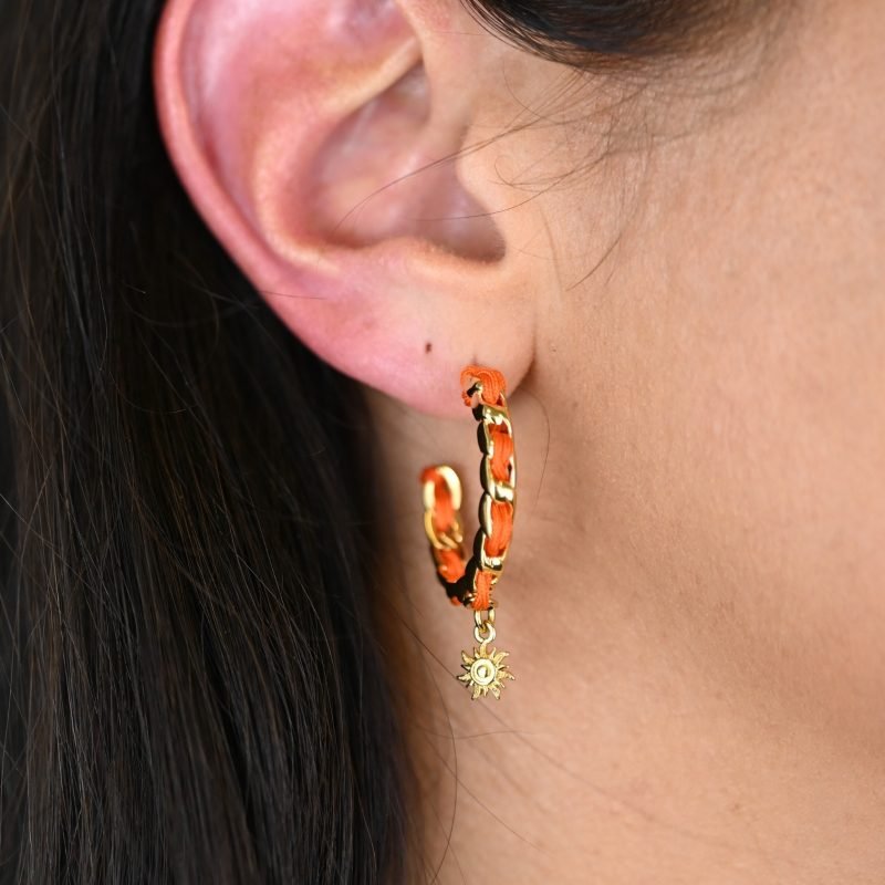 "Les boucles d'oreilles Monsens, inspirées de la collection Couture, où l'on retrouve un célèbre charm et une couleur dominante dans la maille. Taille : 27 mm de diamètre Composition : plaqué or et fil de coton.. Bijou porté, Crédit GEMITY."